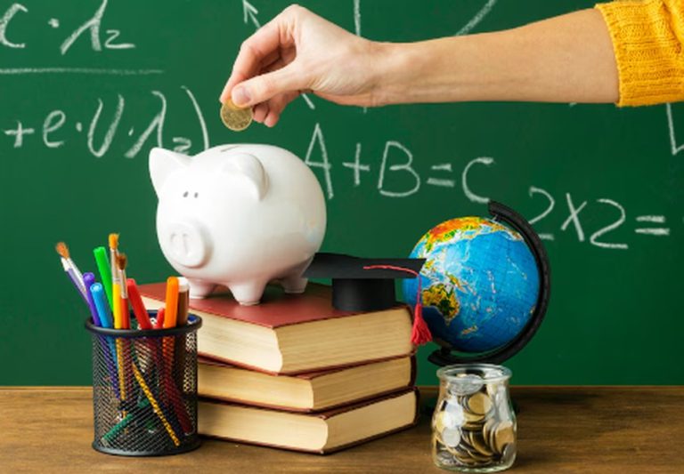 A hand placing a coin into a piggy bank on top of stacked school books, next to a globe, a jar of coins, and school supplies, symbolizing the importance of financial education in schools.