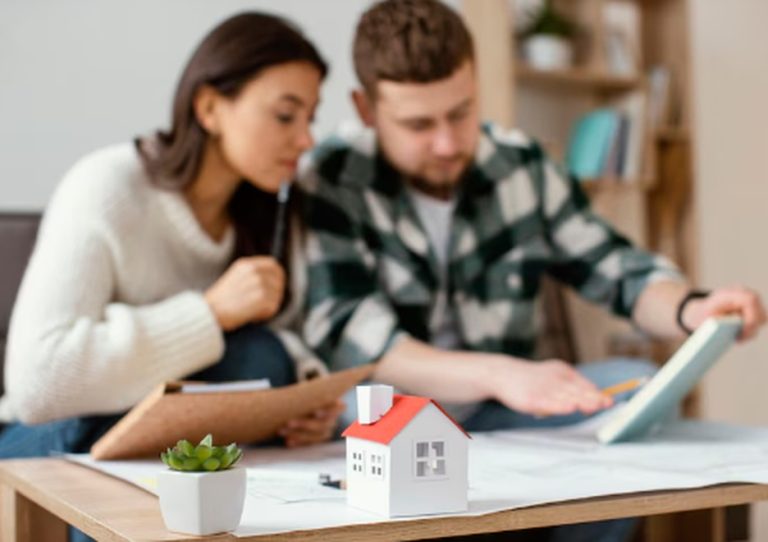 A young couple reviews documents and a digital tablet at home, focusing on financial planning for housing expenses, with a miniature house and paperwork spread out on the table.