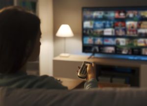 A woman sitting on a couch holding a remote control while browsing multiple streaming services on a smart TV, illustrating the impact of the subscription economy on household entertainment expenses.