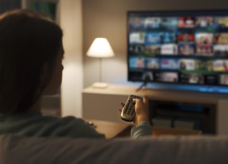 A woman sitting on a couch holding a remote control while browsing multiple streaming services on a smart TV, illustrating the impact of the subscription economy on household entertainment expenses.