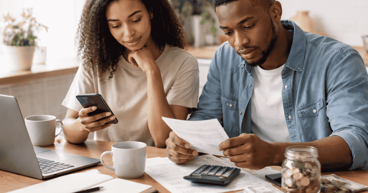 Couple reviewing household finances at a kitchen table, with the man holding a bill and the woman checking information on her smartphone. A laptop, calculator, coffee mugs, and a jar of coins are spread across the table, representing budgeting, expense tracking, and personal financial planning in the United States.
